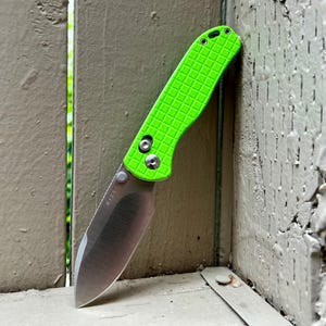 May include: A folding knife with a bright green textured handle and a silver blade. The knife is leaning against a weathered, painted surface. The blade is partially open, showcasing its sharp edge.