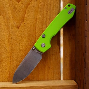 May include: A folding pocket knife with a bright lime green handle and a metallic silver drop point blade. The handle features a textured circular button, a pivot screw, and a small metal pocket clip attached to the top corner of the frame.