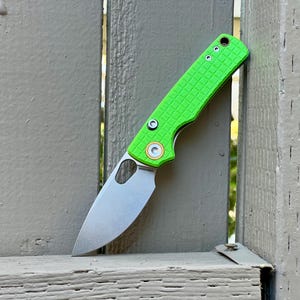 May include: A lime green folding knife with a textured handle and a silver blade. The knife has a circular cutout near the blade's base and a gold-colored accent around the pivot. The handle features three small holes near the top.