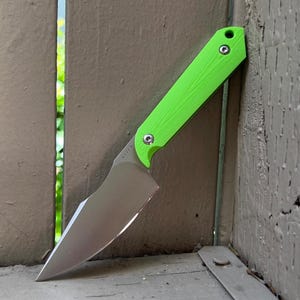 May include: A bright green folding knife with a silver blade. The handle is a vibrant green, and the blade is angled. The knife is leaning against a weathered wooden surface, showcasing its design and craftsmanship.