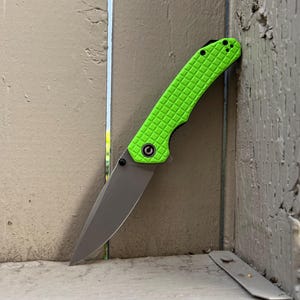 May include: A green pocket knife with a textured handle and a silver blade. The knife is closed and resting against a wooden fence.