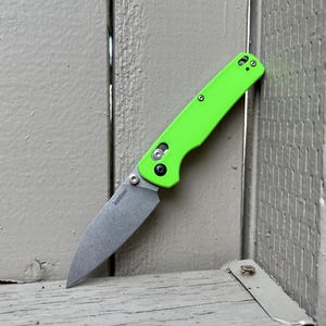 May include: A neon green folding knife with a silver blade. The knife is open, displaying the blade. The handle has a textured surface and visible screws. The brand name "Kershaw" is printed on the blade. The knife is leaning against a weathered wooden surface.