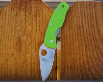 May include: A folding pocket knife with a bright lime green textured handle and a silver stainless steel blade. The blade features a circular thumb hole and a small spider logo engraving near the base of the metal edge.