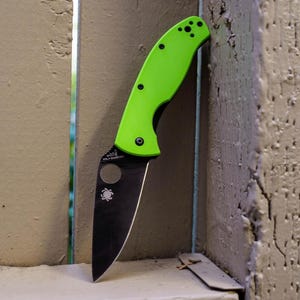 May include: A folding knife with a bright green handle and a black blade. The handle has black hardware and a pocket clip. The blade has a circular cutout and a spider logo. The knife is leaning against a weathered wooden structure.