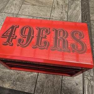 May include: A red toolbox with the word "49ERS" in dark brown lettering across the top. The toolbox has multiple drawers and sits on a tiled surface.