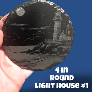May include: A round, dark grey slate coaster featuring an etched lighthouse scene. The design includes a lighthouse, ocean waves, a moon, and the text "4 IN ROUND LIGHT HOUSE #1". The coaster is approximately 10 cm in diameter.
