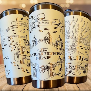 Puede incluir: Tres vasos aislados de color crema con bordes y tapas doradas. Cada vaso presenta un diseño diferente del Mapa del Merodeador, que incluye texto e ilustraciones de Hogwarts. El texto incluye frases como "Juro solemnemente que mis intenciones no son buenas."