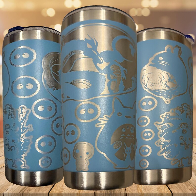 K&ouml;nnte beinhalten: Drei hellblaue Edelstahlbecher mit silbernen Deckeln, die jeweils lasergravierte Illustrationen verschiedener animierter Figuren zeigen, darunter eine gro&szlig;e Kreatur, ein Drache, kleine runde Geister und eine burgartige Struktur auf der Oberfl&auml;che.