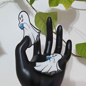 May include: Two white ghost-shaped enamel pins, each holding a blue, multi-sided die. The pins have black outlines and are displayed on a black hand-shaped sculpture. Green leaves are visible in the background.