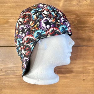May include: A colorful welding cap featuring a repeating pattern of cartoon characters in shades of red, blue, purple, and yellow on a white background. The cap is displayed on a white mannequin head against a wooden background.