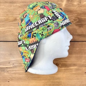 May include: A green and black Minecraft-themed welding cap. The cap features a repeating pattern of Minecraft characters and logos on a green background. The cap has a brim and a neck cover for protection.