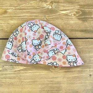 May include: Pink and white fabric hat with a Hello Kitty print. The hat has a curved brim and a pointed crown.
