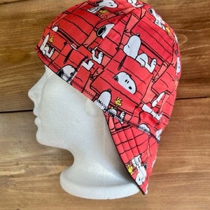 May include: Red welding cap featuring a repeating pattern of Snoopy and his doghouse. The cap is made of a durable fabric and has a curved brim and a sewn seam. The design includes white, black, and yellow accents.
