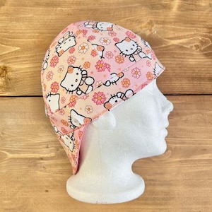 May include: A pink and white welding cap with a Hello Kitty print. The cap has a flap that covers the neck and ears.