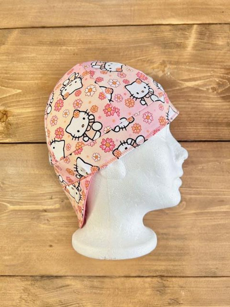 May include: A pink and white welding cap with a Hello Kitty print. The cap has a flap that covers the neck and ears.