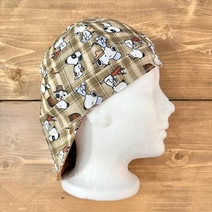 May include: A brown and tan plaid welding cap featuring a repeating pattern of Snoopy in various poses. The cap is made of fabric and has a curved brim. The cap is displayed on a white mannequin head.