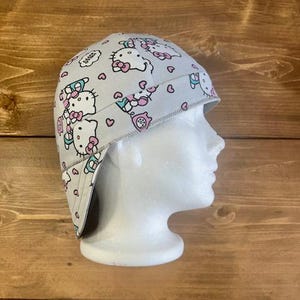 May include: A gray and white welding cap with a Hello Kitty print. The cap has a flap that can be worn down to protect the neck.