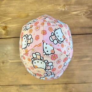 May include: A pink fabric hat with a floral pattern and Hello Kitty characters. The hat is made of a soft, comfortable fabric and is perfect for everyday wear.
