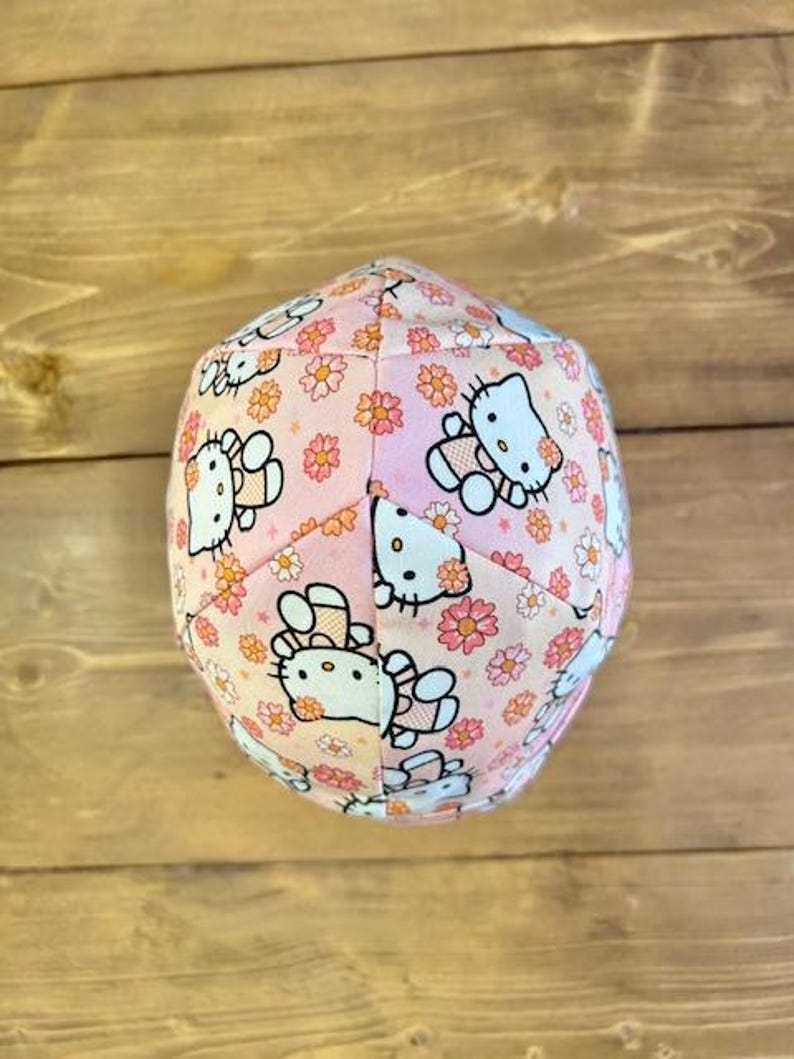 May include: A pink fabric hat with a floral pattern and Hello Kitty characters. The hat is made of a soft, comfortable fabric and is perfect for everyday wear.
