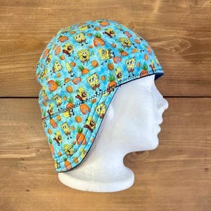 May include: A blue welding cap featuring a repeating pattern of the cartoon character SpongeBob SquarePants, pineapples, and carrots. The cap has a dark blue trim and is displayed on a white mannequin head.