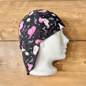 May include: A black scrub cap featuring a repeating pattern of pink cowboy hats, white stars, and grey paisley swirls. The fabric head covering has a fitted crown and a wide band that wraps around the head, providing full coverage for the hair.