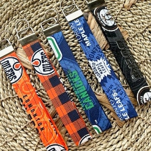 May include: Six different keychains with various NHL team logos. The keychains are made of fabric and feature the team's colors and logos. The keychains are displayed on a woven surface.