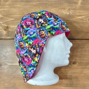 May include: A colorful welding cap featuring a vibrant pattern of cartoon characters in shades of blue, pink, green, and yellow. The cap is displayed on a white mannequin head, showcasing its design and shape. The cap has a pink trim.
