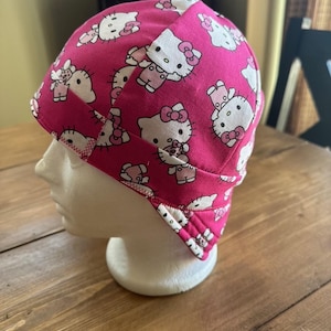 May include: A pink welding cap featuring a repeating pattern of white Hello Kitty characters. The cap is made of fabric and has a white zigzag stitch along the edge. The cap is displayed on a white mannequin head.