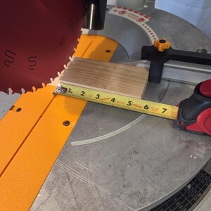 May include: A close-up of a miter saw with a wooden block being measured. The tape measure shows the block is approximately 6 inches long. The saw blade is red and the saw base is silver and orange.