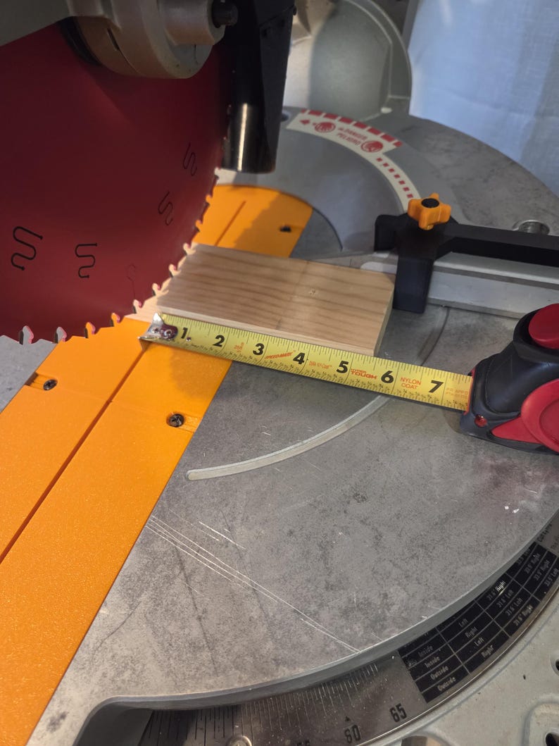 May include: A close-up of a miter saw with a wooden block being measured. The tape measure shows the block is approximately 6 inches long. The saw blade is red and the saw base is silver and orange.
