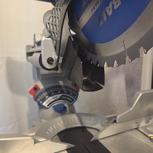 Op de afbeelding: Een Kobalt verstekzaag met een blauwe en zilveren afwerking, een cirkelzaagblad met 40 tanden en een schaalverdeling voor dubbele afschuining. De metalen voet bevat een hoekindicator en de zaagkop bevindt zich boven het vlakke werkoppervlak.