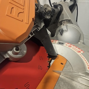 K&ouml;nnte beinhalten: Nahaufnahme einer silbernen und orangefarbenen Gehrungss&auml;ge. Das S&auml;geblatt ist sichtbar, mit einem roten und orangefarbenen Blattschutz. Die S&auml;ge hat einen schwarzen Griff und einen silbernen Sockel. Der Markenname "RIDGID" ist auf dem orangefarbenen Geh&auml;use zu sehen.