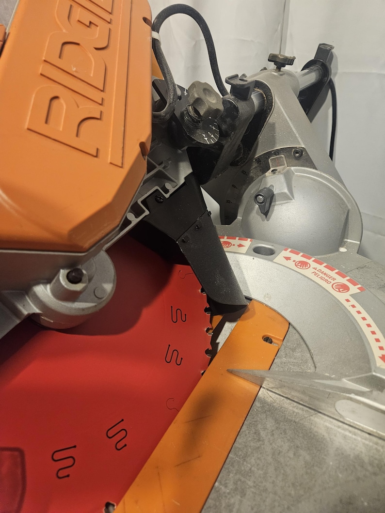 K&ouml;nnte beinhalten: Nahaufnahme einer silbernen und orangefarbenen Gehrungss&auml;ge. Das S&auml;geblatt ist sichtbar, mit einem roten und orangefarbenen Blattschutz. Die S&auml;ge hat einen schwarzen Griff und einen silbernen Sockel. Der Markenname "RIDGID" ist auf dem orangefarbenen Geh&auml;use zu sehen.