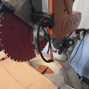 May include: A Ridgid brand miter saw with a red blade and orange housing. Safety glasses hang from the saw. A light-colored wooden plank is positioned on the saw's base, ready for cutting. The saw is designed for woodworking projects.