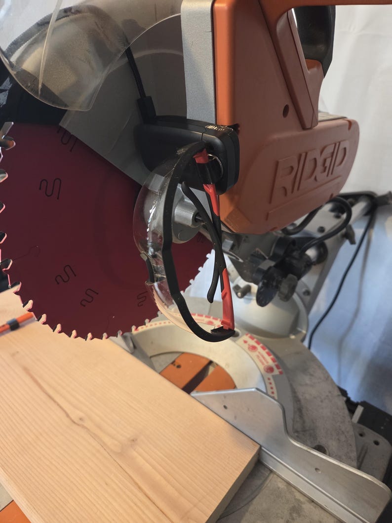 May include: A Ridgid brand miter saw with a red blade and orange housing. Safety glasses hang from the saw. A light-colored wooden plank is positioned on the saw's base, ready for cutting. The saw is designed for woodworking projects.