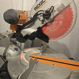 K&ouml;nnte beinhalten: Eine silberne und orangefarbene Ridgid-Gehrungss&auml;ge mit einem gro&szlig;en, roten Kreiss&auml;geblatt. Die S&auml;ge hat einen transparenten Klingenschutz und einen schwarzen Griff. Das Ridgid-Logo ist auf der Seite der S&auml;ge zu sehen.