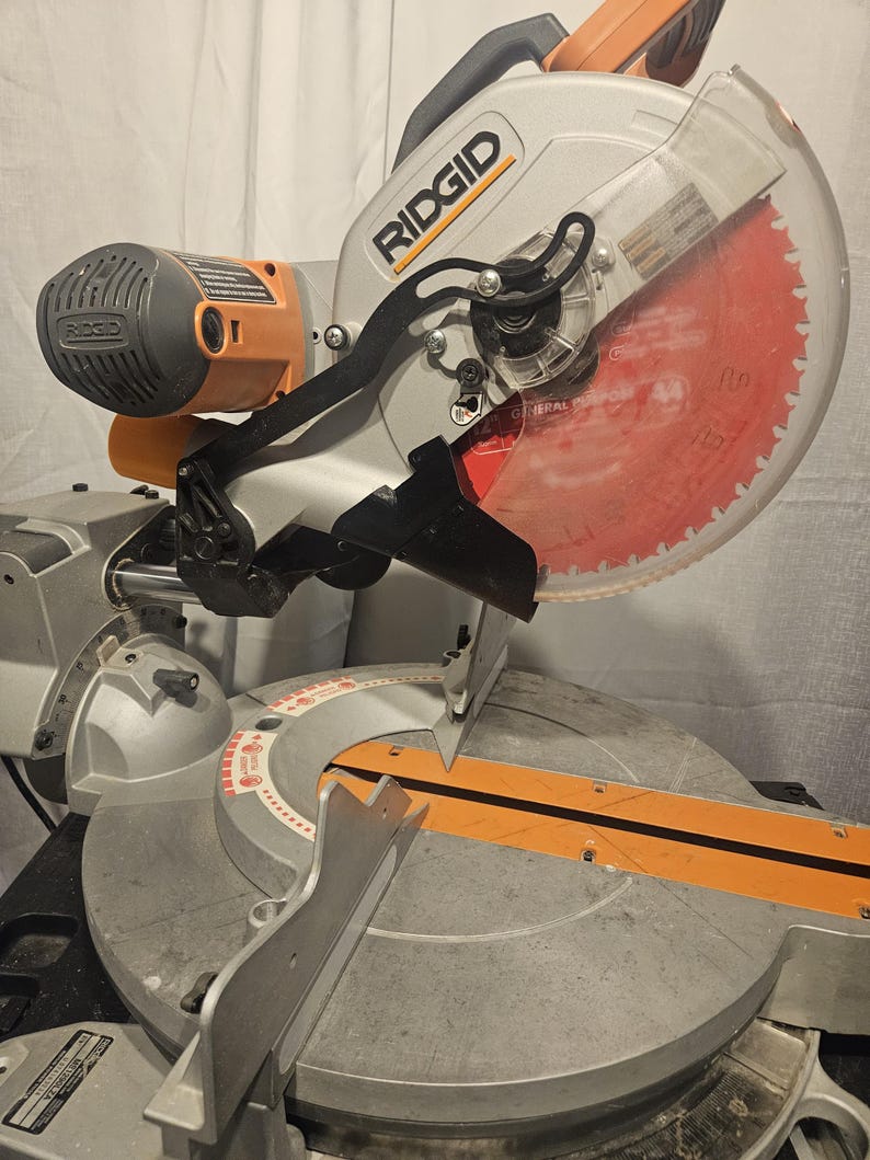 K&ouml;nnte beinhalten: Eine silberne und orangefarbene Ridgid-Gehrungss&auml;ge mit einem gro&szlig;en, roten Kreiss&auml;geblatt. Die S&auml;ge hat einen transparenten Klingenschutz und einen schwarzen Griff. Das Ridgid-Logo ist auf der Seite der S&auml;ge zu sehen.