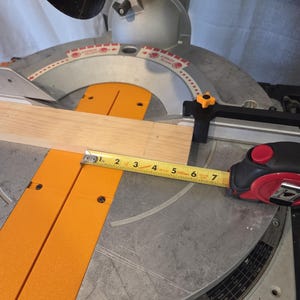 May include: A miter saw with a wooden plank and a measuring tape. The saw has a red blade and orange supports. The tape measure shows a measurement of approximately 6 inches.