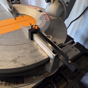May include: A close-up of a miter saw with a black and orange blade guard. The saw has a silver base and an orange table. The blade is silver and angled. The saw is on a light-colored surface.