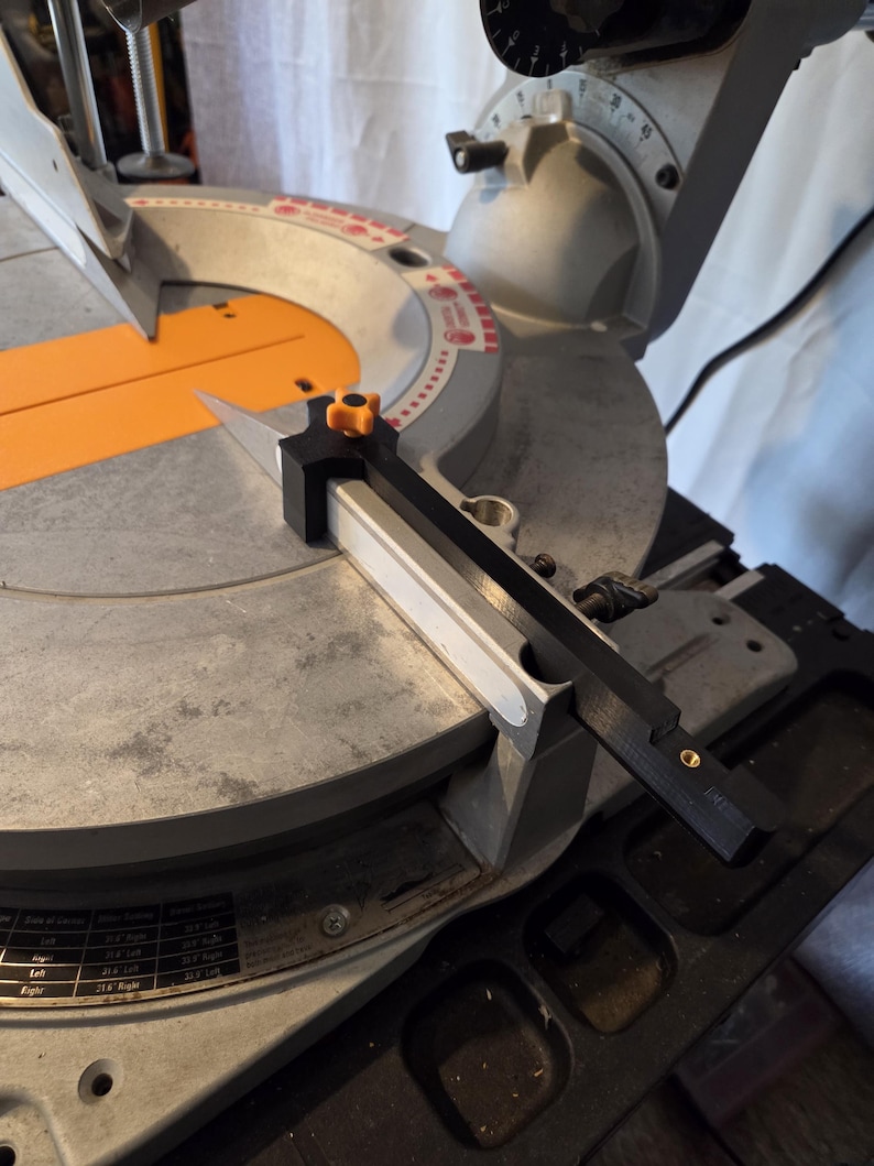 May include: A close-up of a miter saw with a black and orange blade guard. The saw has a silver base and an orange table. The blade is silver and angled. The saw is on a light-colored surface.