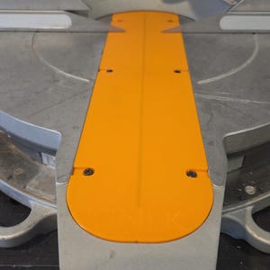 May include: Close-up of a miter saw with a bright orange blade guard and a silver metal base. The saw has a circular blade and a scale for precise angle cuts. The design is industrial and functional.