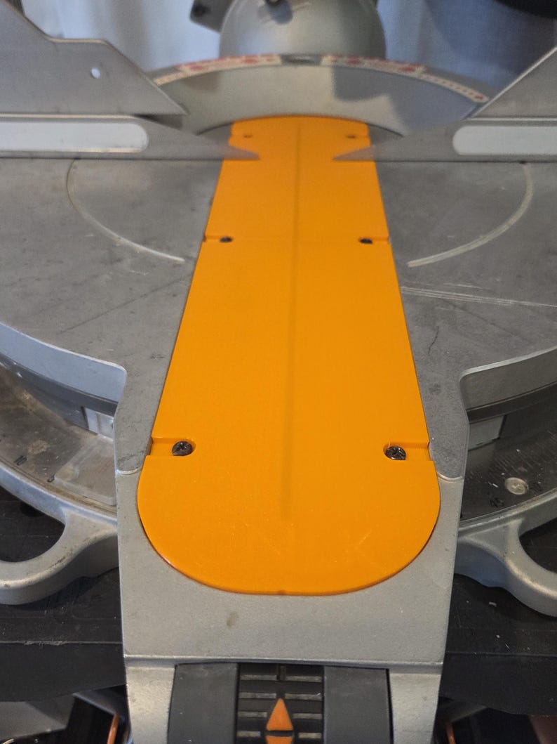 May include: Close-up of a miter saw with a bright orange blade guard and a silver metal base. The saw has a circular blade and a scale for precise angle cuts. The design is industrial and functional.