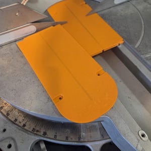 May include: Close-up of a miter saw with two bright orange blade guards. The saw has a silver blade and a gray base with degree markings. The guards are rectangular with rounded ends and small holes.