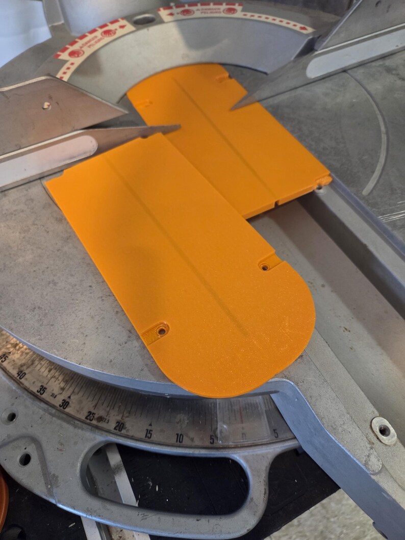 May include: Close-up of a miter saw with two bright orange blade guards. The saw has a silver blade and a gray base with degree markings. The guards are rectangular with rounded ends and small holes.