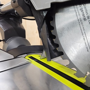 May include: Close-up of a grey and black mitre saw with a yellow cutting guide. The saw blade is visible and has a maximum operating speed of 5,000 RPM.