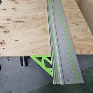 May include: A silver Festool guide rail with green markings. The rail is lying on a piece of plywood.