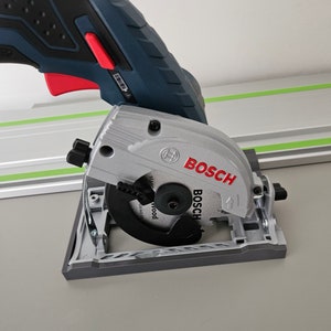May include: A silver and black Bosch circular saw with a red trigger. The saw blade is black and the handle is dark blue. The word "BOSCH" is printed in red on the blade guard. The saw is designed for cutting wood.