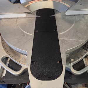 May include: A silver and black miter saw with a black base and a circular blade. The saw has a black handle and a silver blade guard. The base has the letters "GNLK" printed on it.