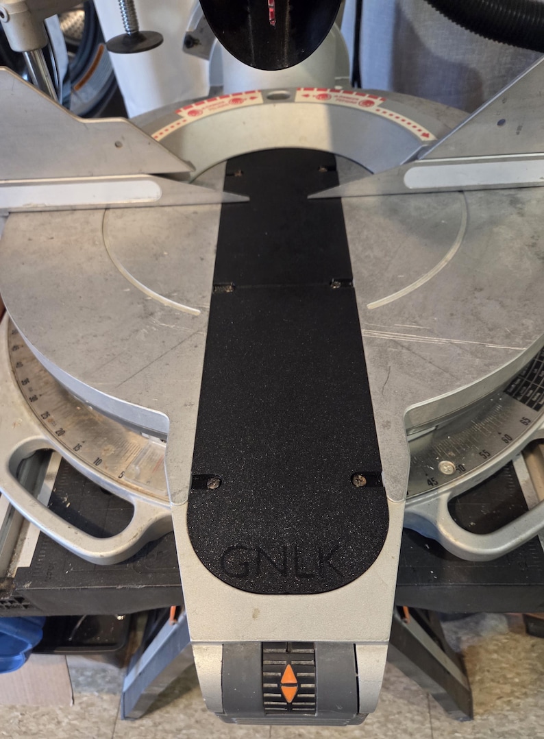 May include: A silver and black miter saw with a black base and a circular blade. The saw has a black handle and a silver blade guard. The base has the letters "GNLK" printed on it.