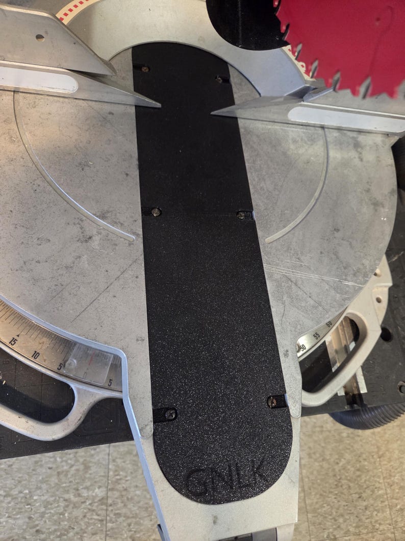May include: A close-up of a miter saw with a black textured surface and a silver blade. The saw has a measurement scale and the letters "GNLK" are visible on the base.